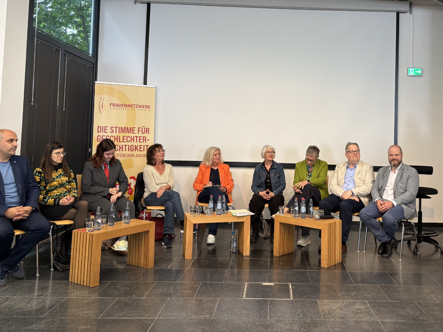 Auf dem Foto sitzt Solveig Münstermann zusammen mit anderen Menschen auf einer Bühne. Im Hintergrund steht ein Rollup vom Frauennetzwerk StädteRegion Aachen.